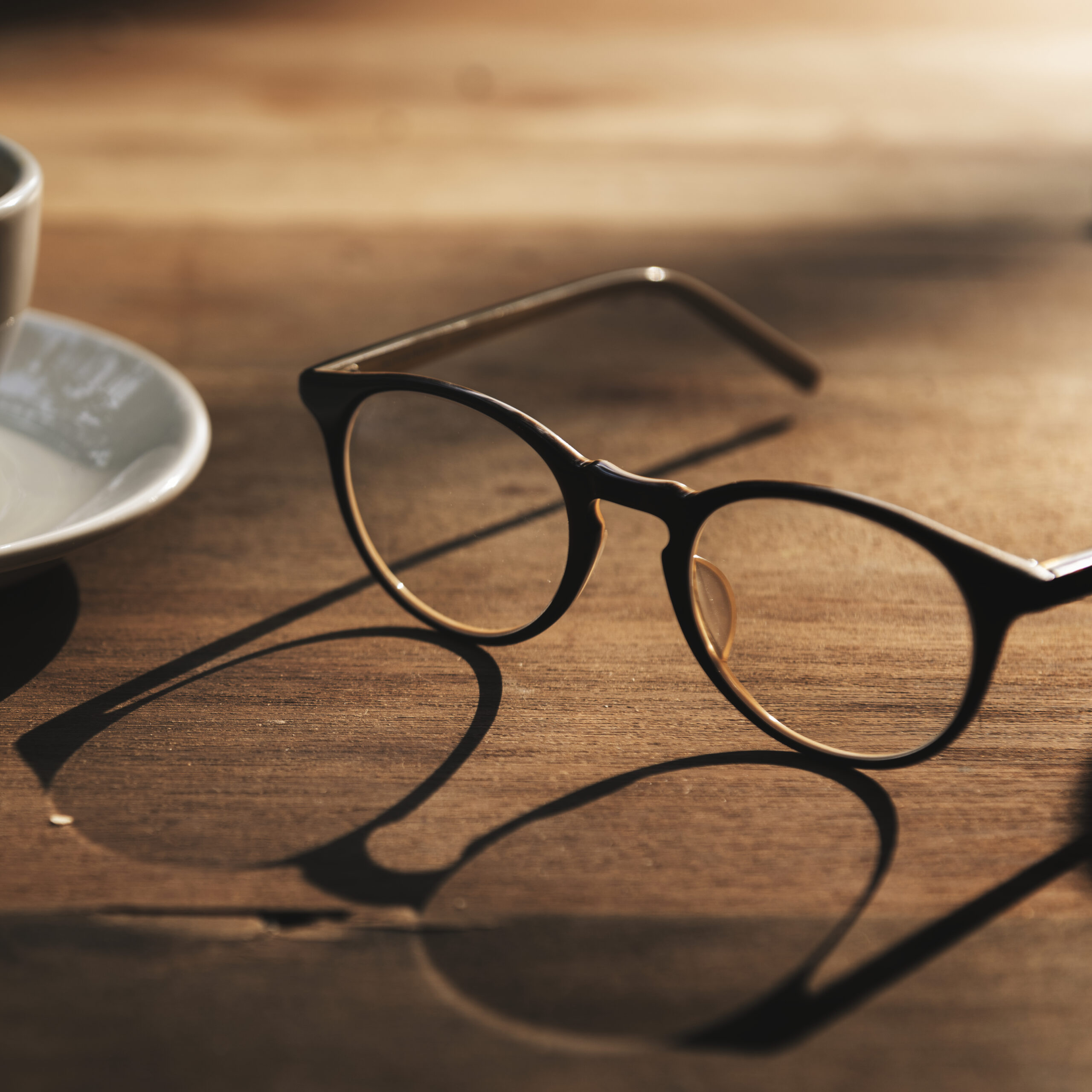 Armação de óculos preta sobre mesa de madeira ao lado de uma xícara de café